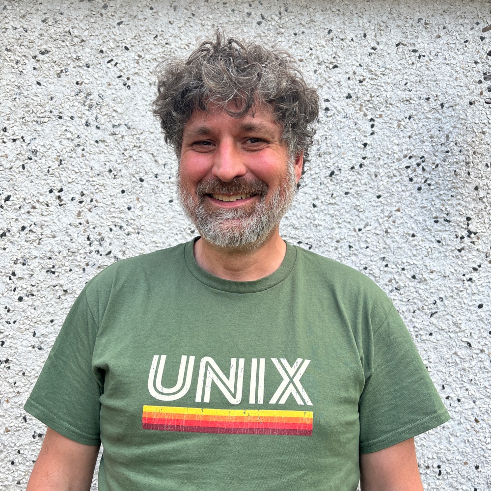 A man in his 50s, brown-grey hair and beard, smiling. He's wearing a T-shirt with "UNIX" printed on the chest.